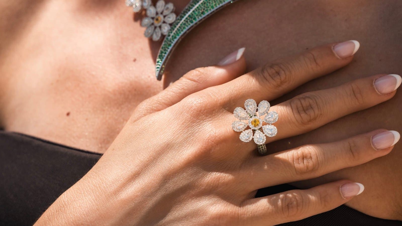 A close-up of a woman's hand wearing a diamond flower ring, with a diamond and emerald necklace visible on her chest.